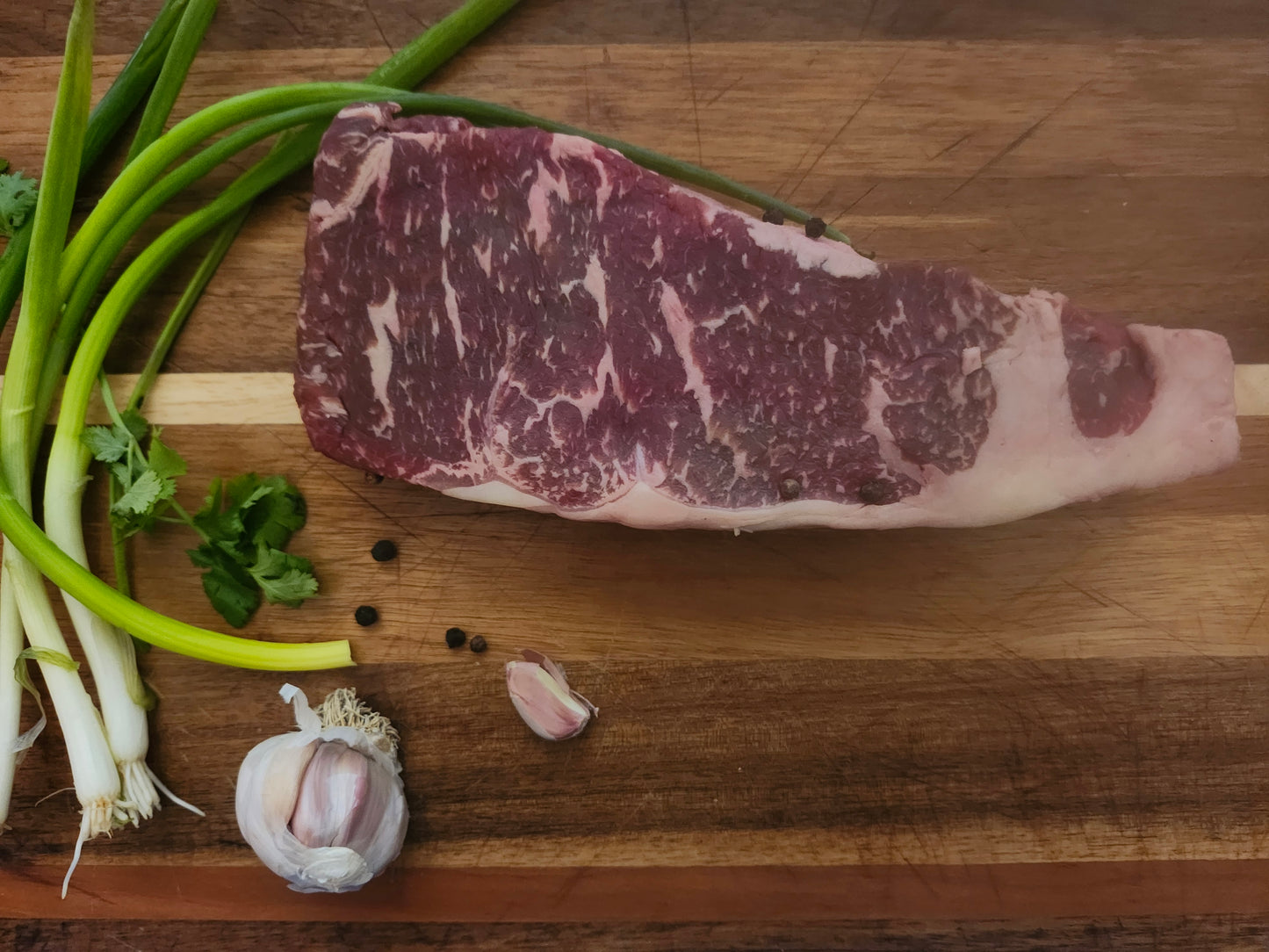 American Wagyu NY strip steak on cutting board with green onion parsley garlic and peppercorn 